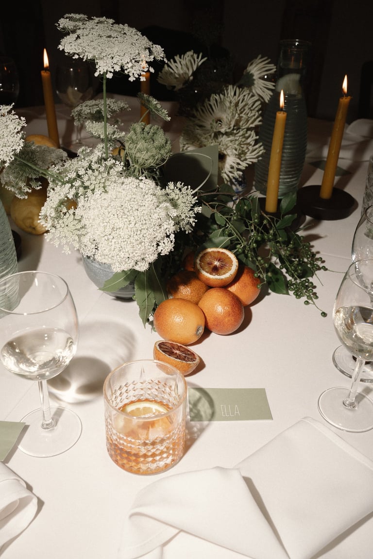 Elegantiškai dekoruotas stalas su baltomis gėlėmis, žalumynais, raudonais apelsinais ir kokteiliu