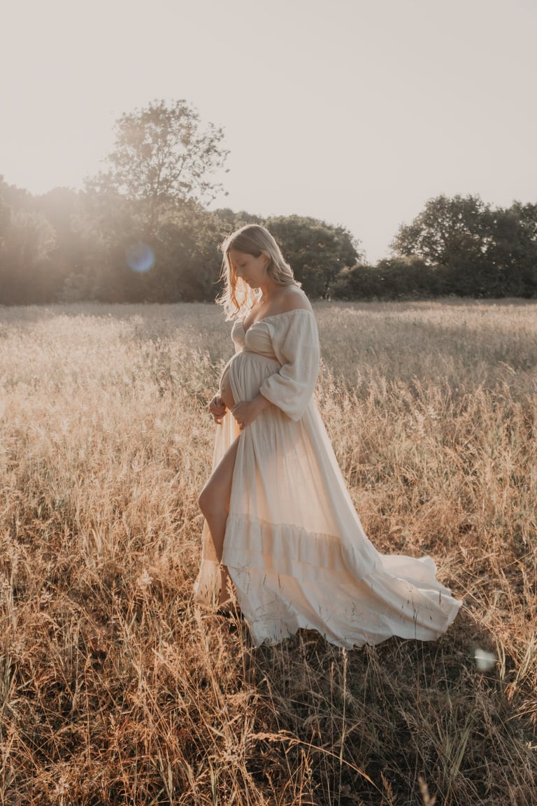 A pregnant woman in a flowing maternity dress poses in a golden sunlit meadow for an outdoor maternity photoshoot.