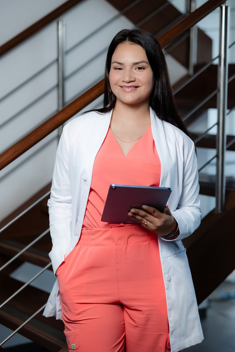 Female healthcare professional in a white lab coat and orange scrubs holding a digital tablet.