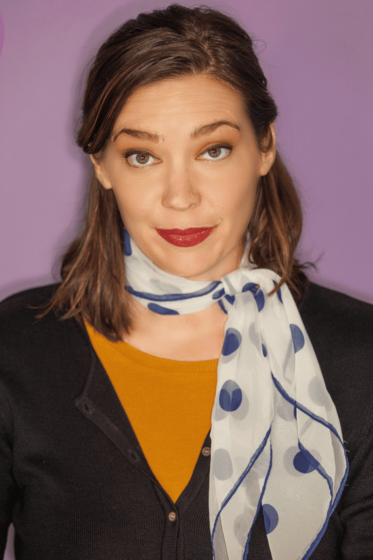 A smiling woman with brown hair and a white and blue polka dot scarf, wearing a mustard shirt and black cardigan, against a