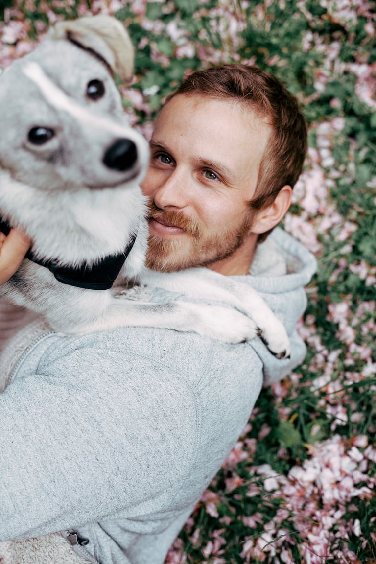 Porträt eines Mannes, der im Gras mit rosa Blütenblättern liegt und seinen Hund anschaut.