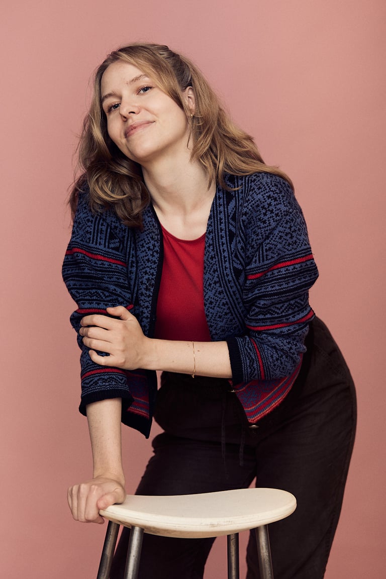 A young woman wearing a blue sweater and red t-shirt leaning on a stool.