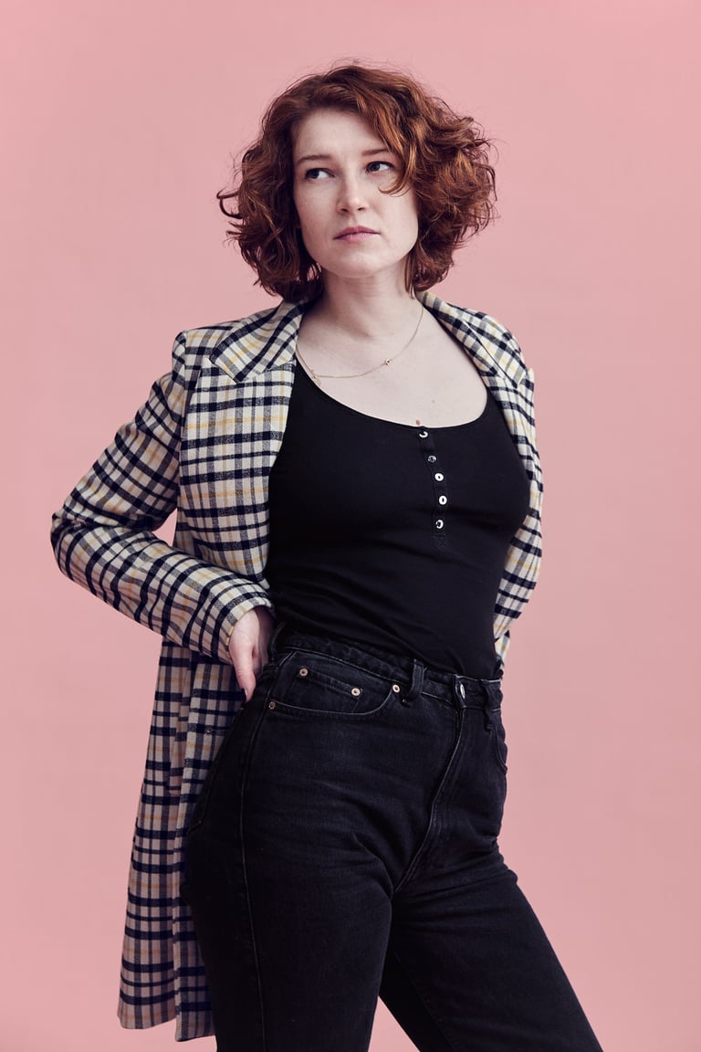 A young woman with red hair standing in front of a pink background.