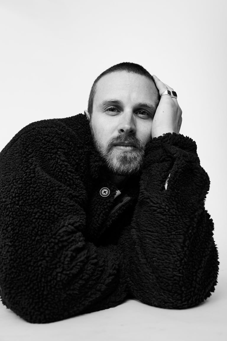 A black and white portrait of a man lying on a floor in a black woollen jacket.
