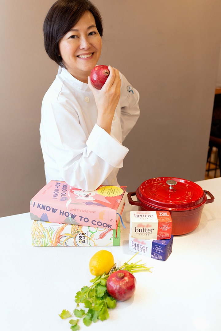 a woman in a chef's uniform holding a red pomegranate smiling at the camera