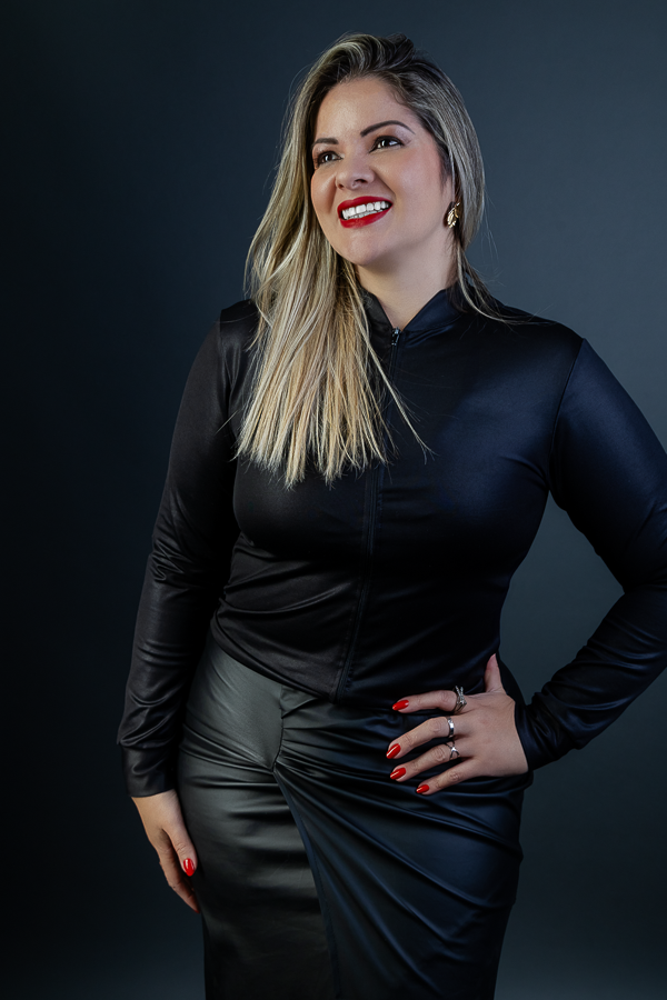 mulher loira vestida de preto com batom vermelho e mão na cintura, sorrindo e olhando ao lado