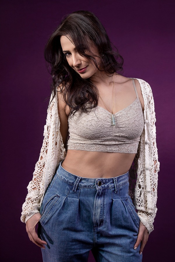 mulher com cabelos ondulados , roupa de crochê caída nos ombros, com leve sorriso em fundo roxo