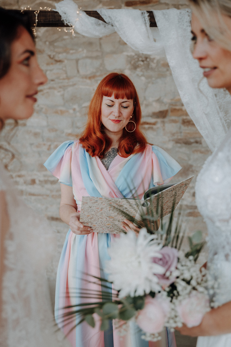 das queere Hochzeitspaar während der Trauung. die zwei Bräute sehen sich liebevoll an. die Rednerin blickt in ihre Rede-Mappe