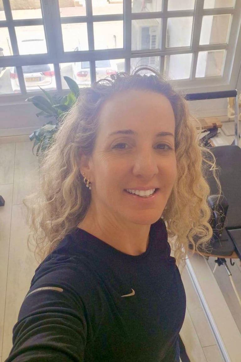 Smiling woman with curly hair wearing activewear in a pilates studio with reformer equipment.