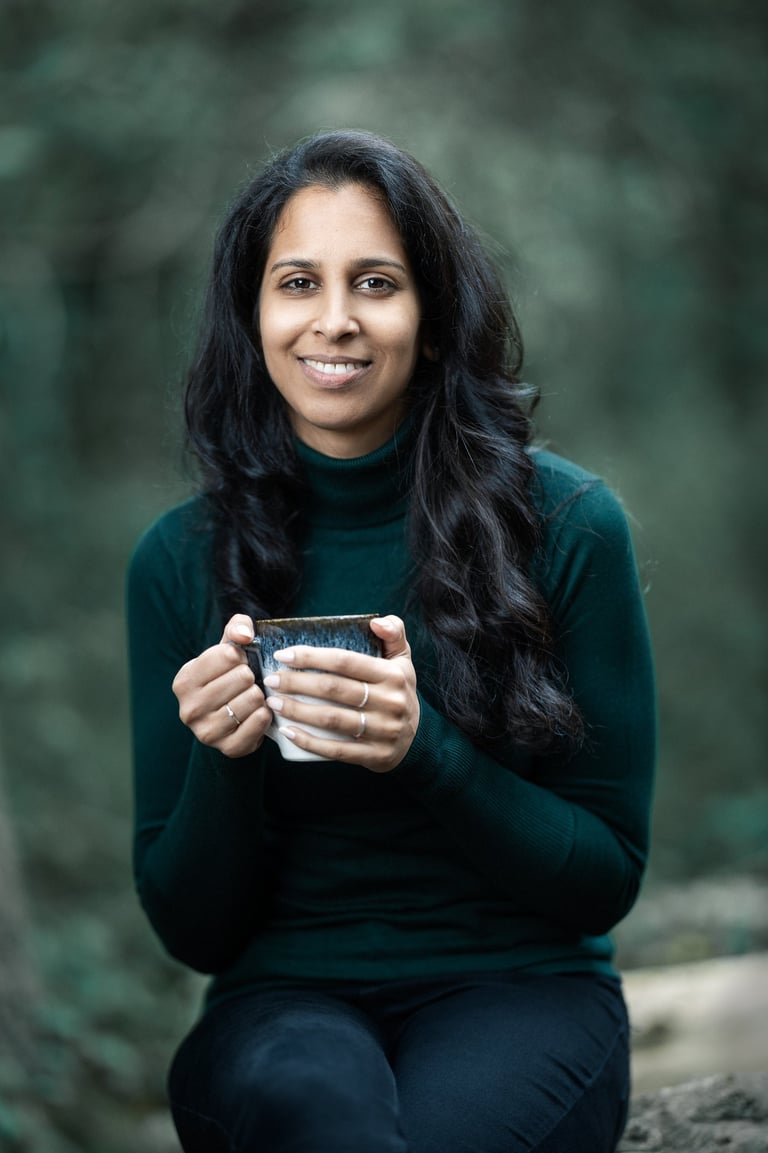 Shivani gently smiling, holding a mug and sitting in a woodland