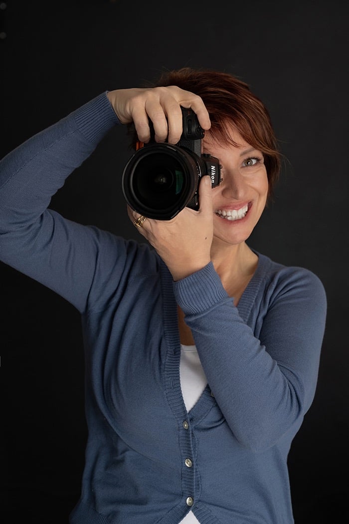 Smiling female professional photographer with red hair holding a Nikon DSLR camera against a black background.
