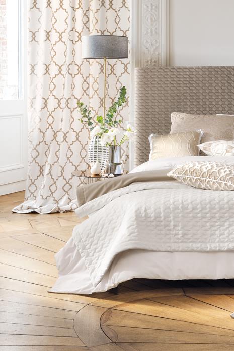 Elegant bedroom with neutral bedding, textured headboard, and pattern curtains on parquet flooring.