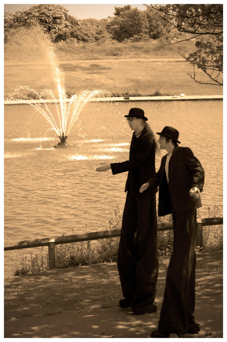 Two street performers on high stilts in black suits walk past a park water fountain.