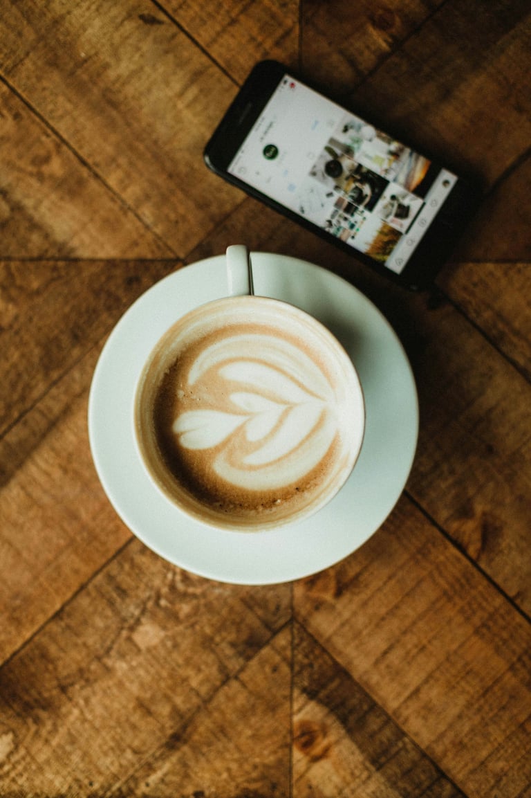 Taza de café con latte art sobre mesa de madera junto a un teléfono móvil, reflejando el ritual y es