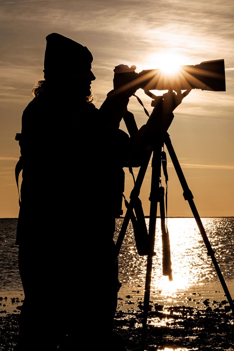 Silhouette of a photographer using a DSLR camera and tripod to capture a golden sunset over the ocean.