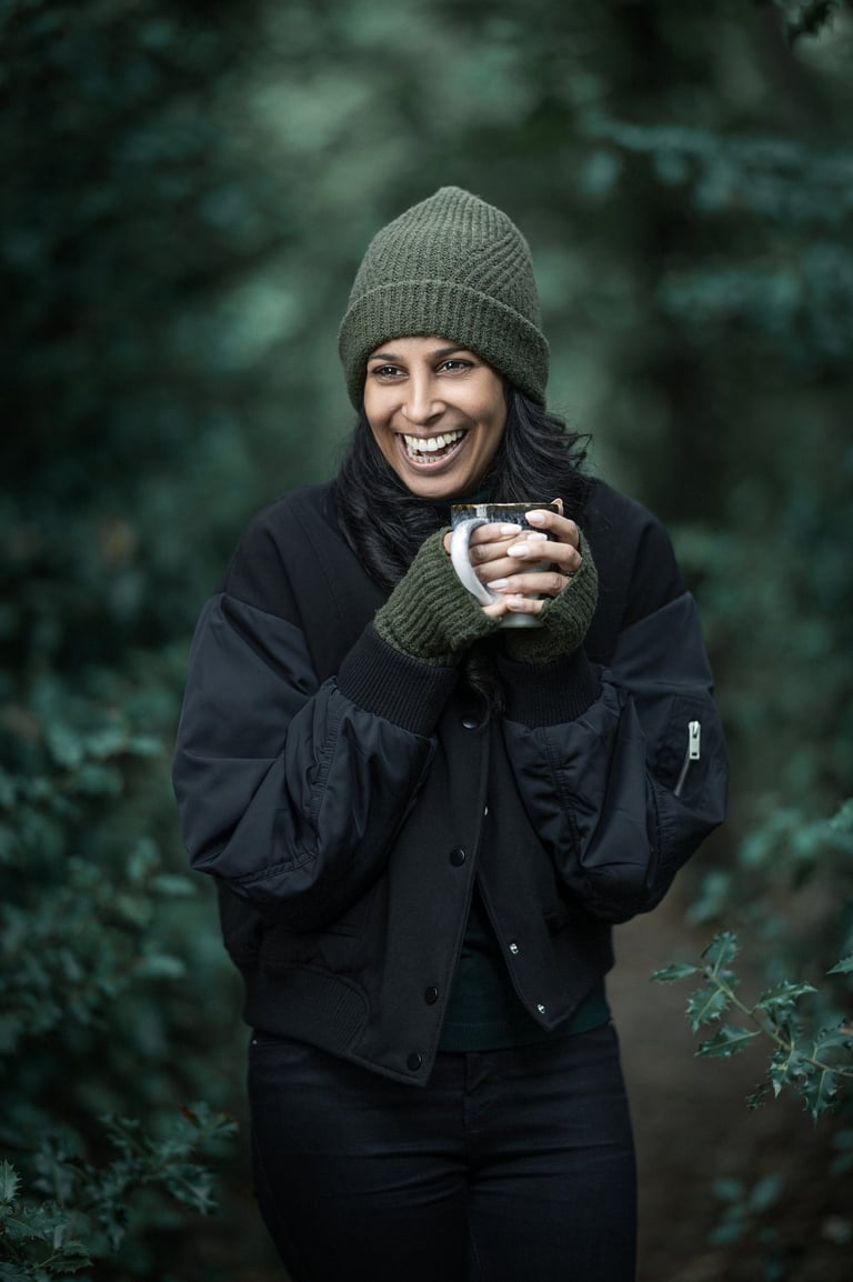 Shivani wearing winter clothes, grinning, holding a mug and standing in woodland.