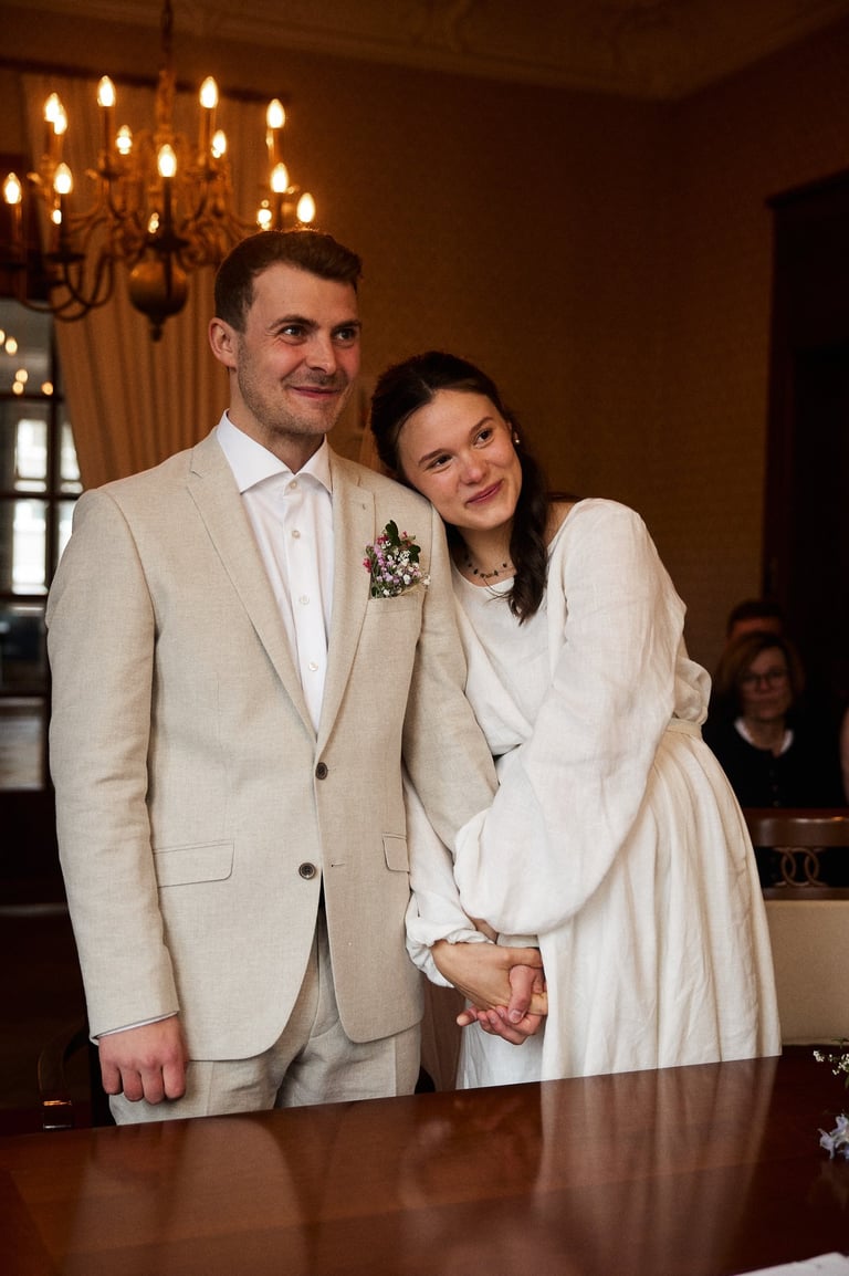 A smiling bride in a white linen dress leans on her groom in a tan suit at their wedding ceremony.