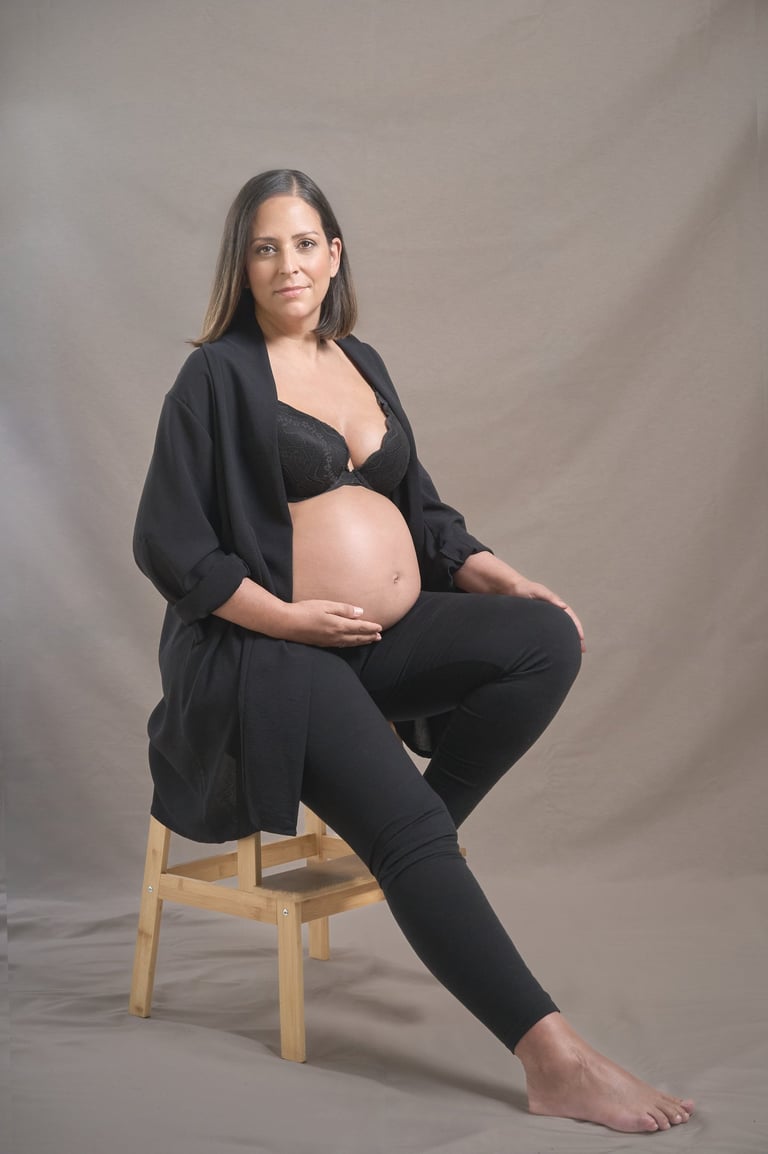 A pregnant woman in a black outfit poses for a maternity photoshoot sitting on a wooden stool.