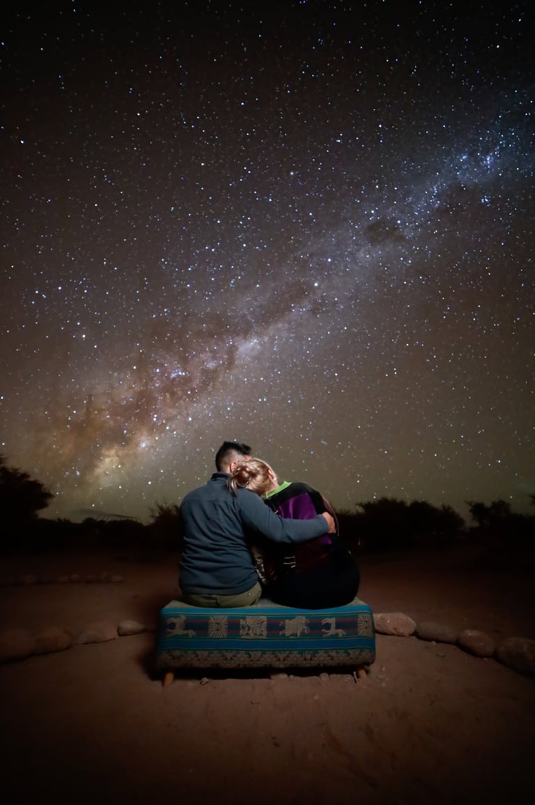 A couple sits on a bench under the vibrant Milky Way galaxy and a clear starry night sky.