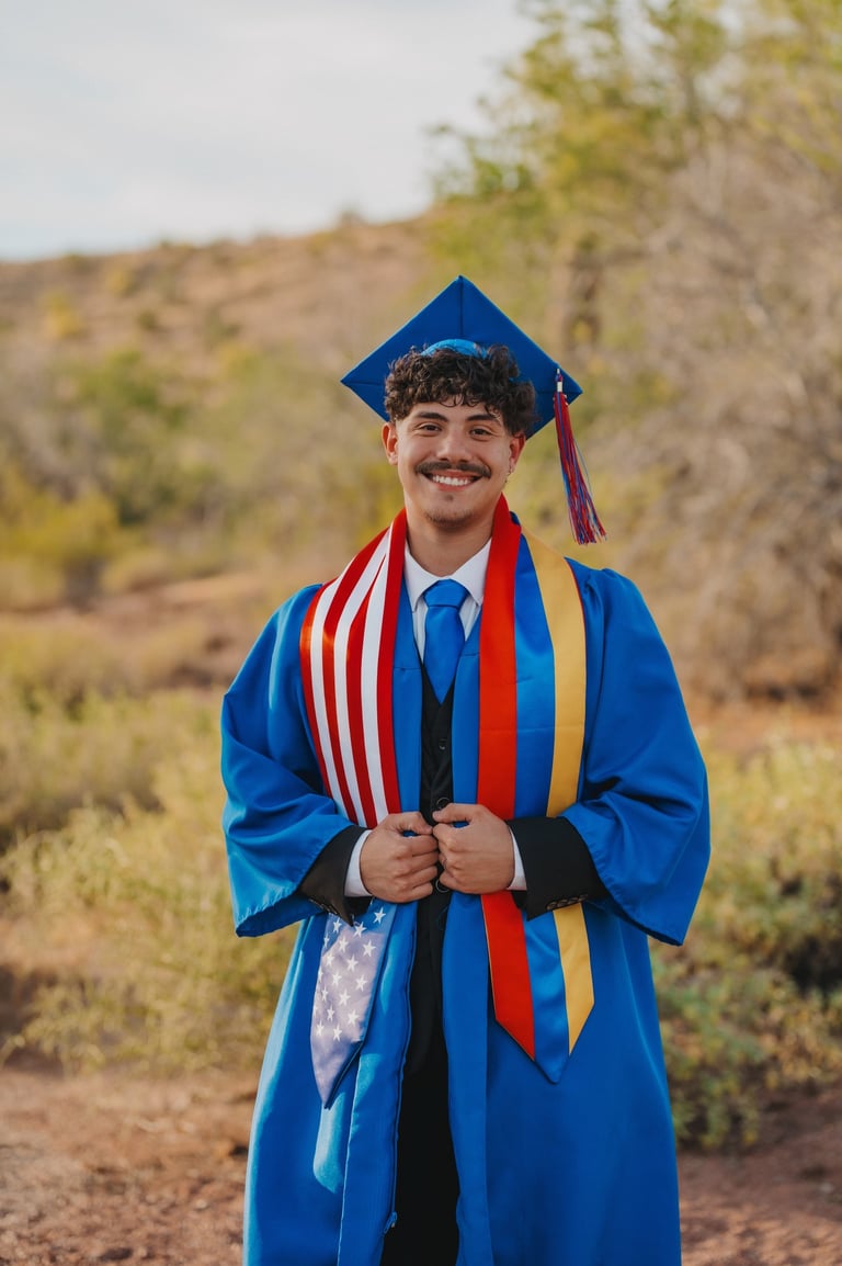 A smiling male graduate in a blue cap and gown wears a patriotic American flag stole for an outdoor portrait.