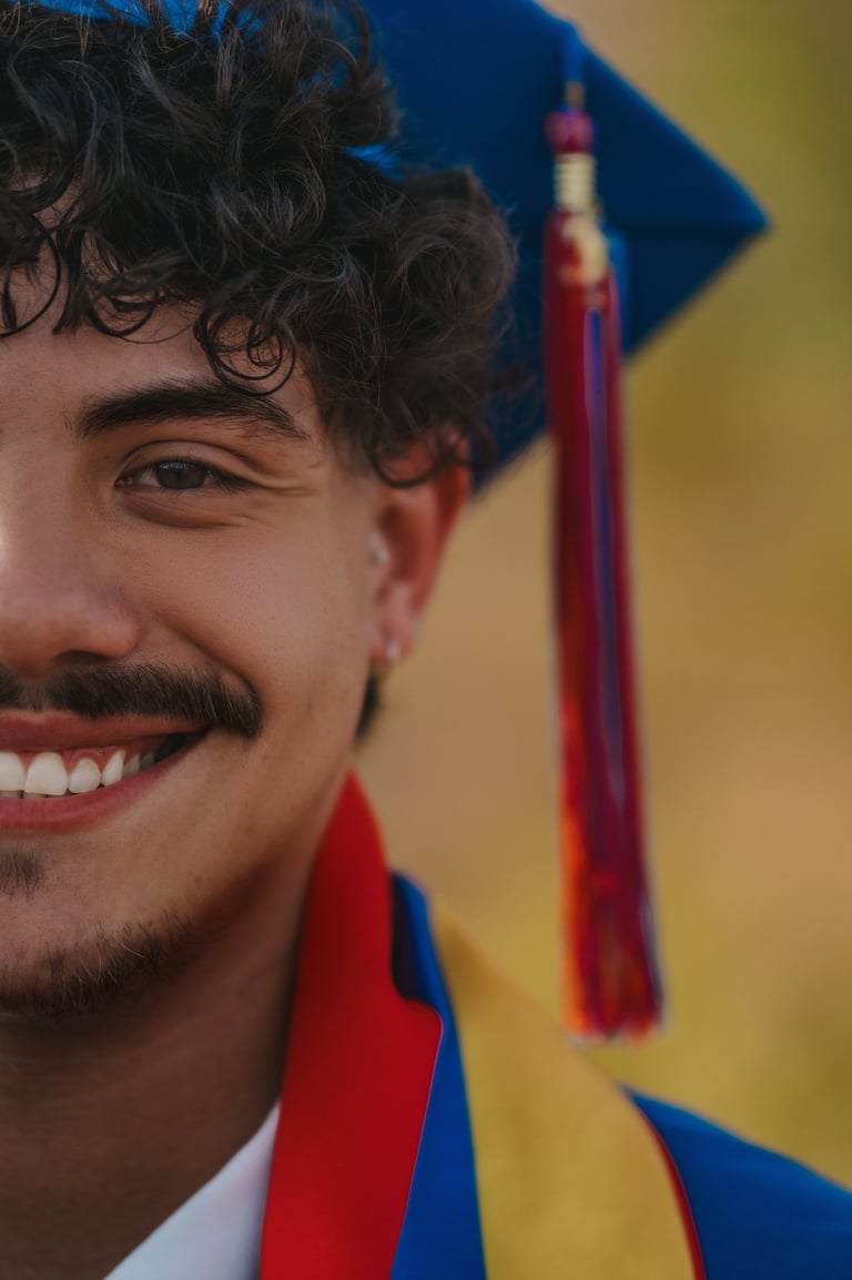 A smiling male graduate with curly hair wearing a blue graduation cap and colorful tassel.