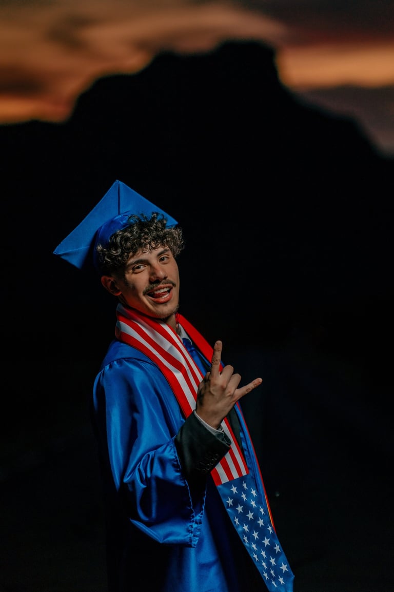 Smiling male graduate in blue cap and gown wearing a US flag stole during a sunset portrait.