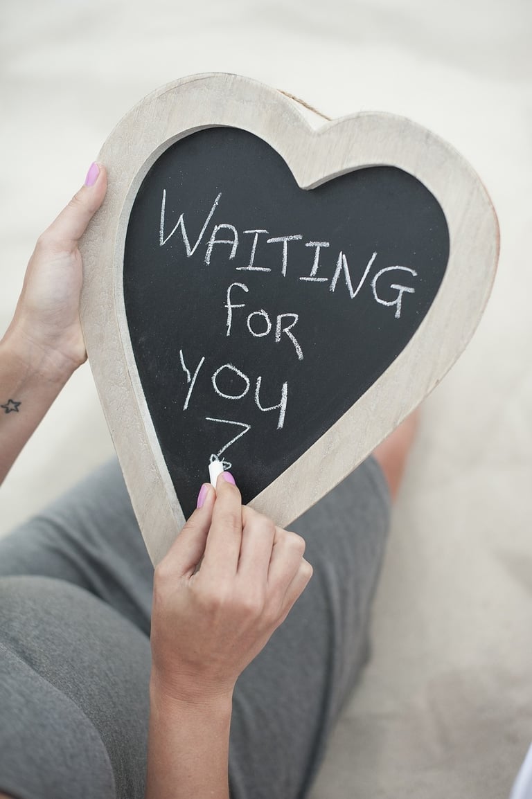 Pregnant woman writing Waiting For You on a heart-shaped chalkboard for a maternity photo shoot.