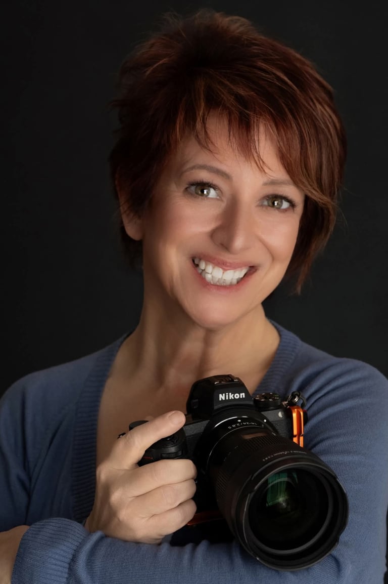 A smiling woman with short auburn hair holding a professional Nikon mirrorless camera.