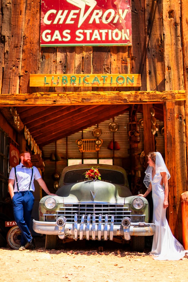 A bride and groom posing with a vintage car at a rustic Chevron gas station wedding venue at Nelson 