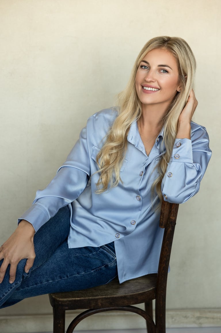 Moteris koučerė sėdi ant rudos kėdės, šypsosi, pasirėmusi ranka galvą ir dėvinti žydrus marškinius