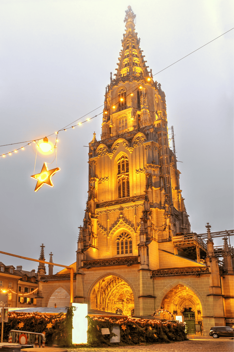 The illuminated Gothic spire of Bern Minster cathedral at dusk with festive Christmas lights and stars.