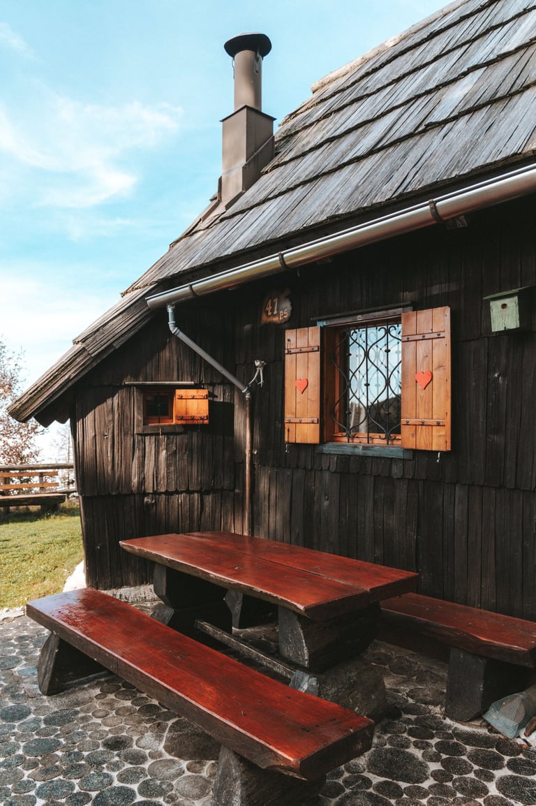 Rustic wooden alpine cabin with heart-carved shutters and an outdoor picnic table on a stone patio.
