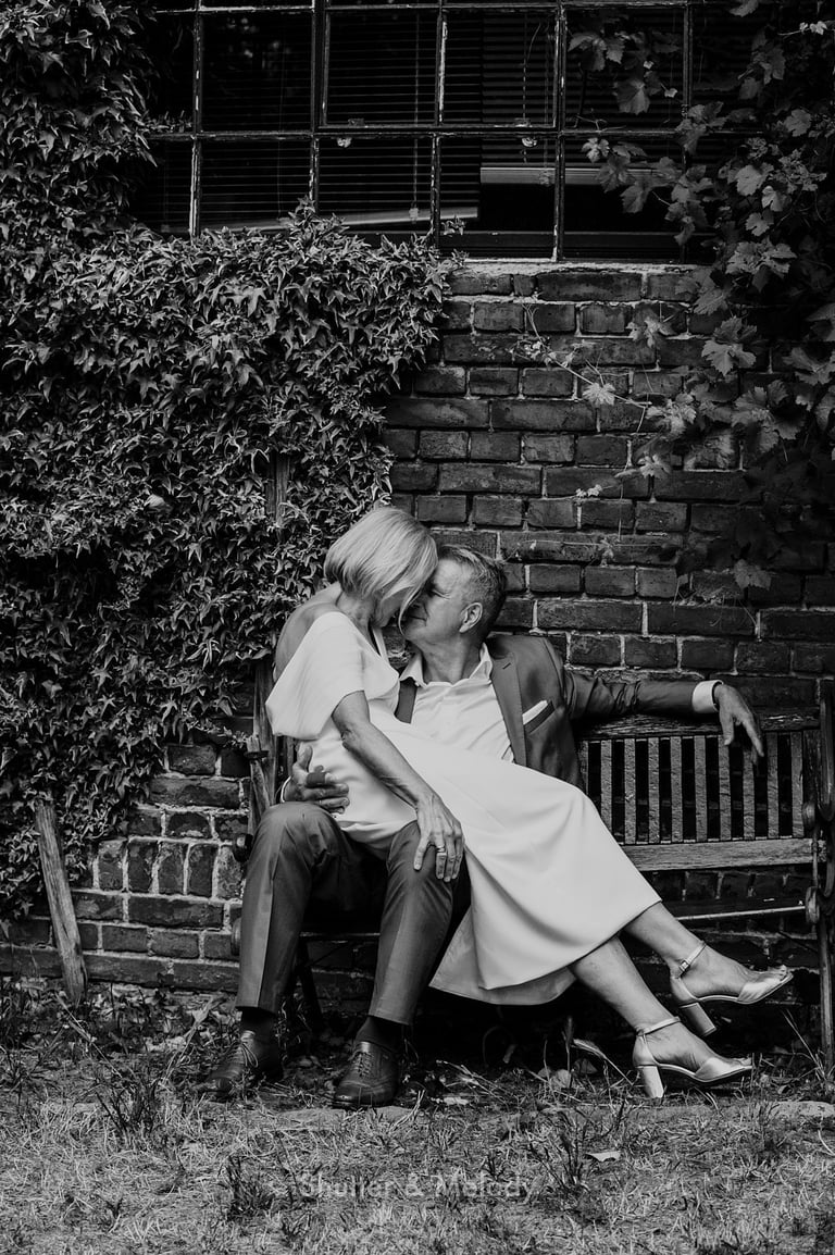 Bride sitting on the groom's knees in front of a brick building.