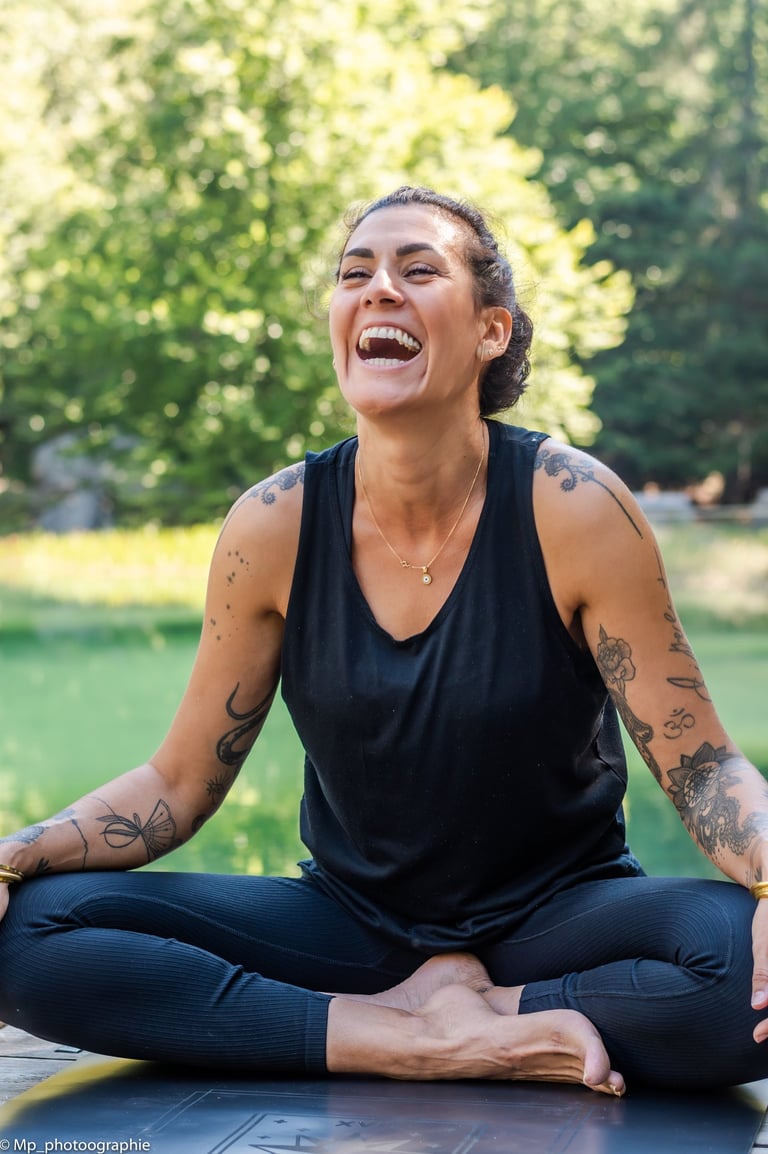 A tattooed woman laughing while practicing outdoor yoga in a cross-legged seated pose.