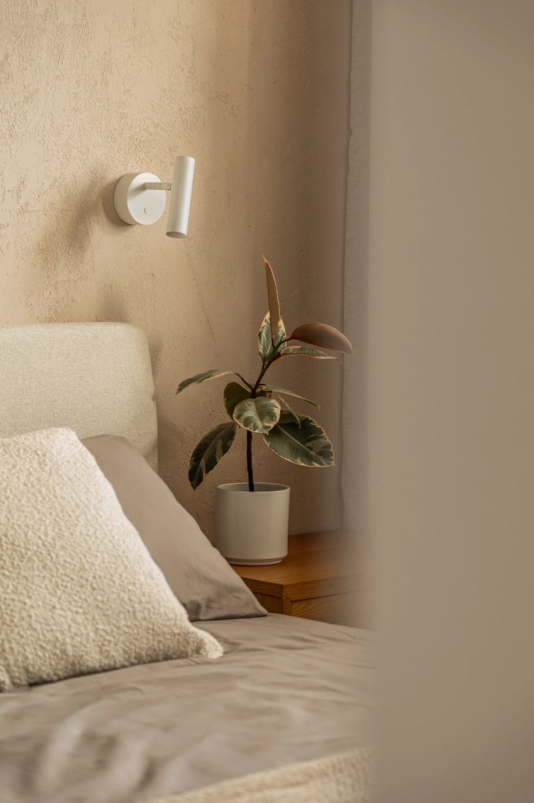 Biały kinkiet do czytania nad łóżkiem i roślina na szafce nocnej w sypialni.