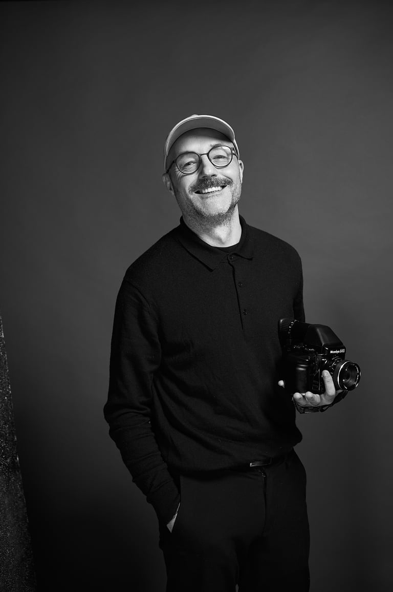 A smiling photographer in a cap holding a professional camera against a grey background.