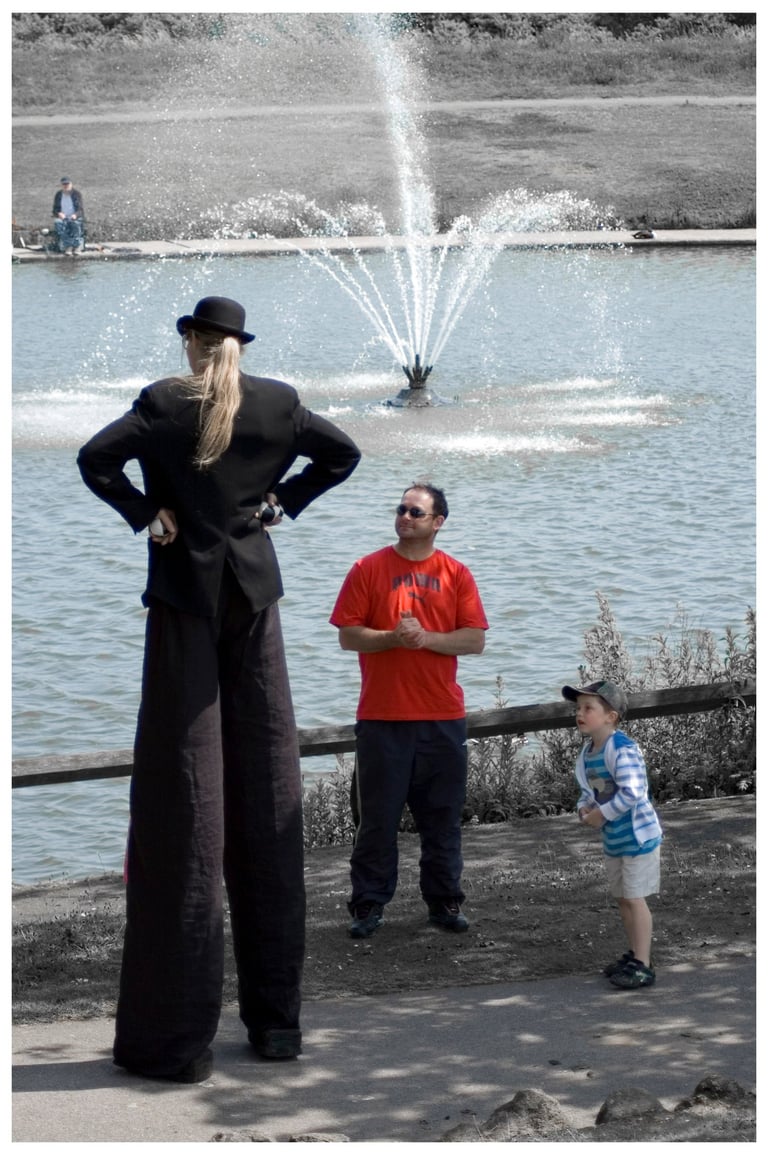 Street performer on tall stilts entertains a man and child by a park fountain.
