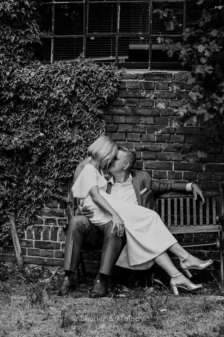 Bride sitting on the groom's knees in front of a brick building.