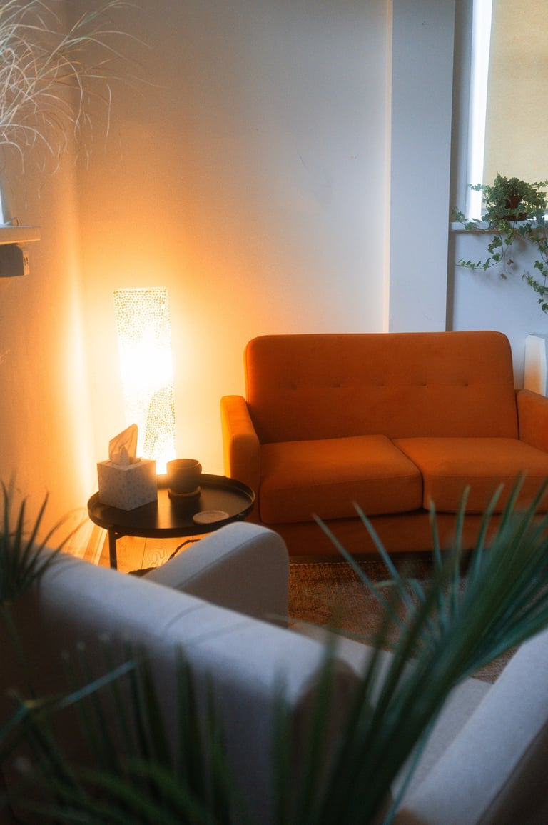 An orange couch sits across from a grey chair. There are plants, a rug, and warm lighting.