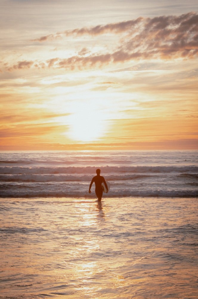 A silhouette of a surfer standing in the ocean, walking into the sun. Vibrant orange and yellow