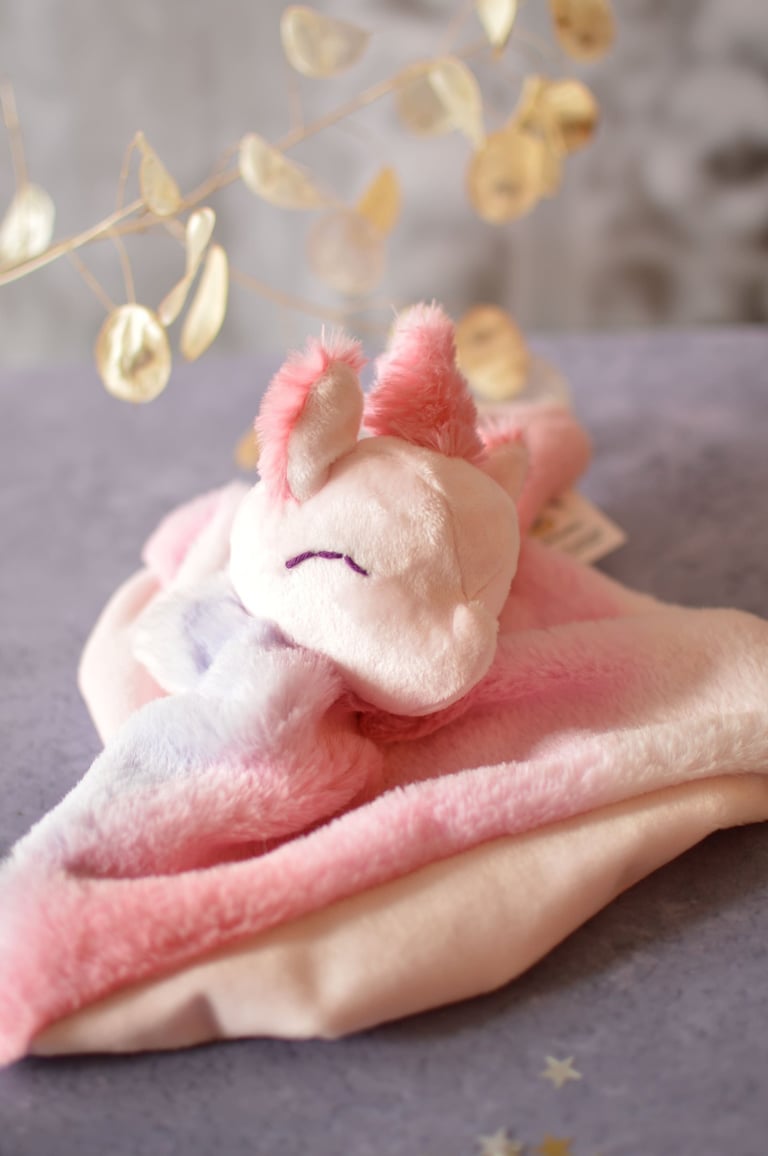 Doudou licorne artisanal en tissu rose pastel, visage brodé, posé dans une ambiance douce.