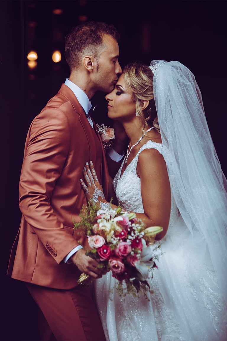 Mariés s'embrassant tendrement : marié en costume terracotta, mariée en robe blanche avec voile.