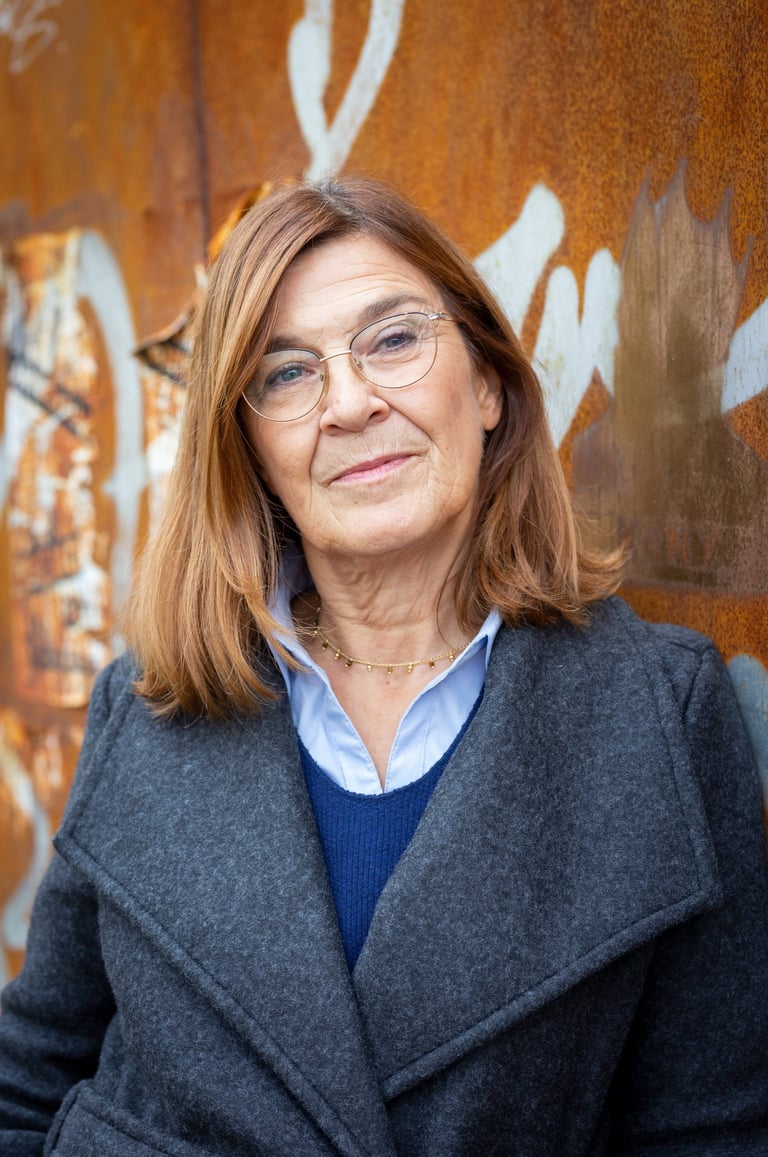 Professional middle-aged woman with glasses and red hair wearing a wool coat against a rusty wall.