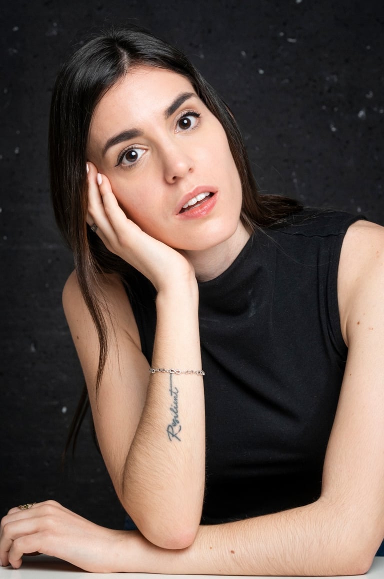 Professional headshot of a young woman with dark hair and a resilient arm tattoo posing against a dark background.
