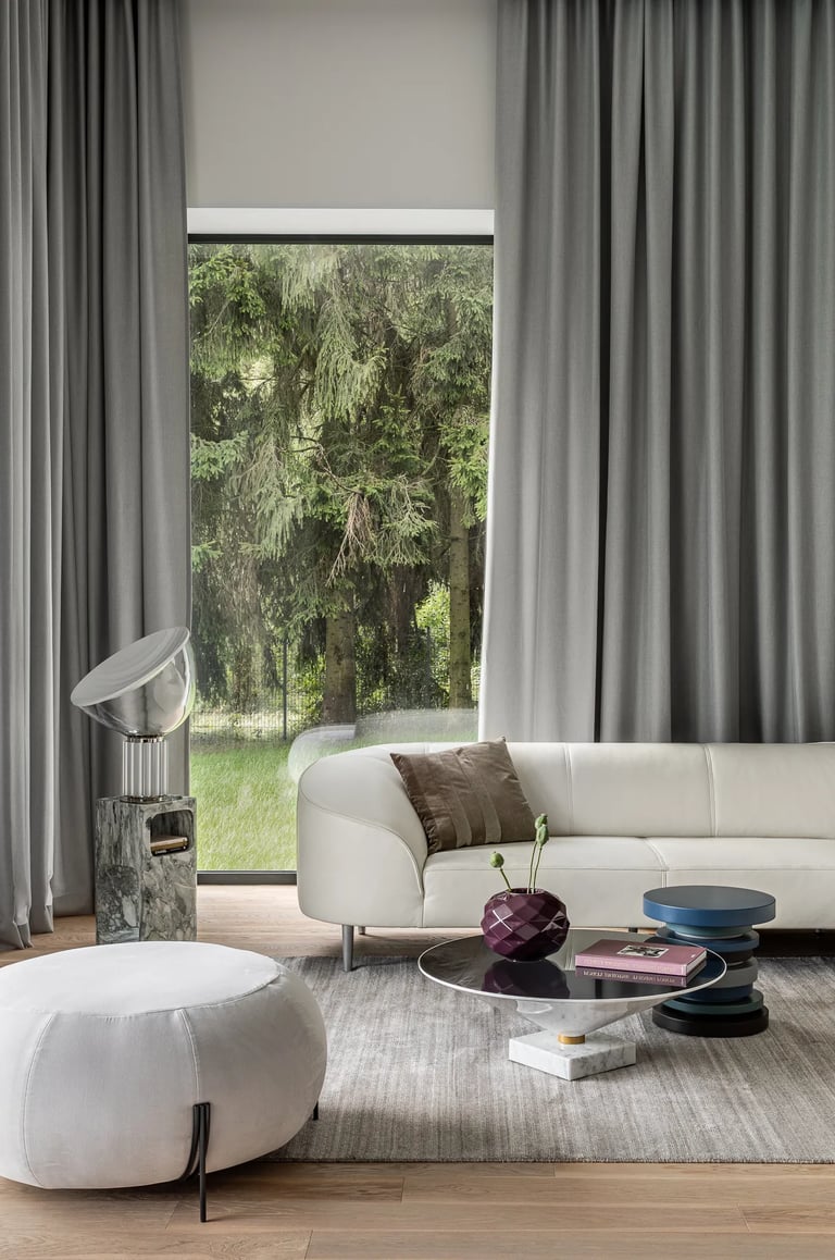 Aranżacja jasnego salonu, biała skórzana sofa, designerskie stoliki kawowe i szare zasłony.