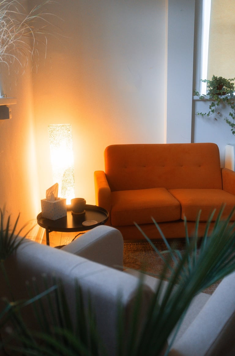 An orange couch sits across from a grey chair. There are plants, a rug, and warm lighting.