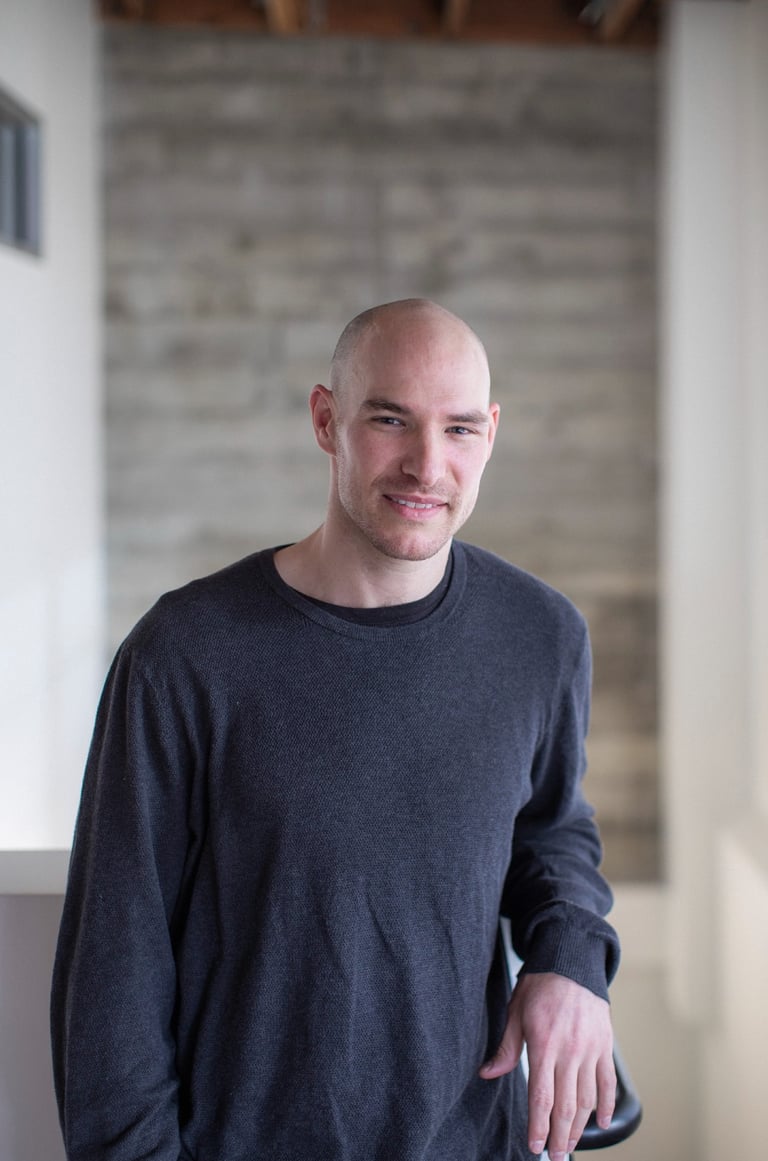 Professional bald man in a casual grey sweater posing for a portrait in a modern office.