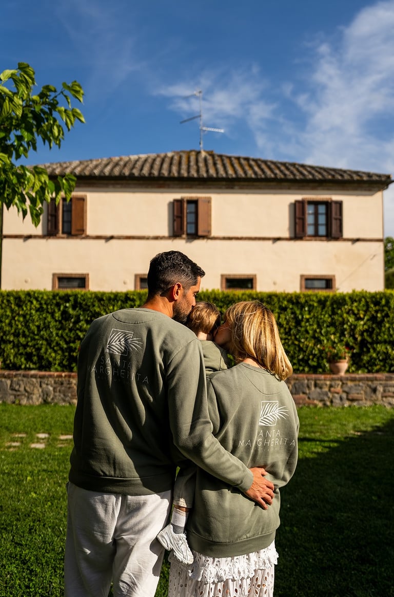 A family wearing matching green sweatshirts hugs in a garden in front of a rustic Italian villa.