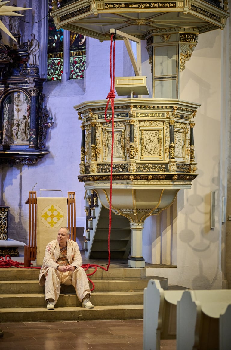Ein Schauspieler sitzt in einer Kirche auf einer Treppe unter der Kanzel. Über ihm ein Galgenknoten.