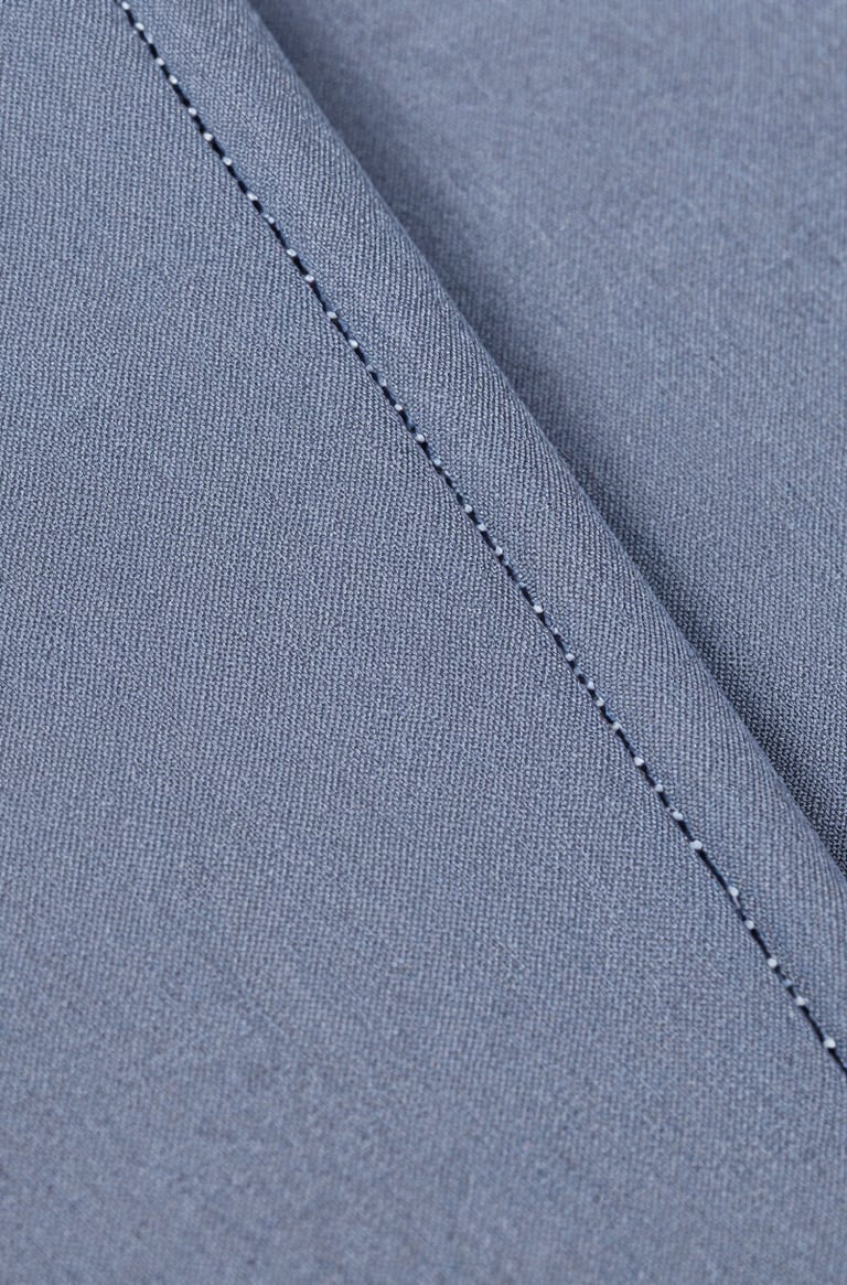 Extreme close-up of school uniform fabric — tight weave of a grey-blue cotton-poly blend, thread interlace fully visible, a reinforced seam running diagonally through the frame, shot under bright even studio light with no shadows, full detail across the surface